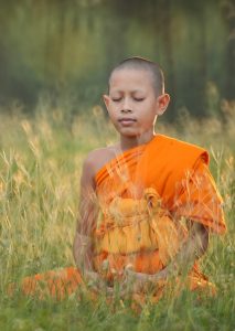 Auf einer Wiese sitzt ein Kind in seinem hell-orangenen, buddhistischen Gewand in seiner Meditation.